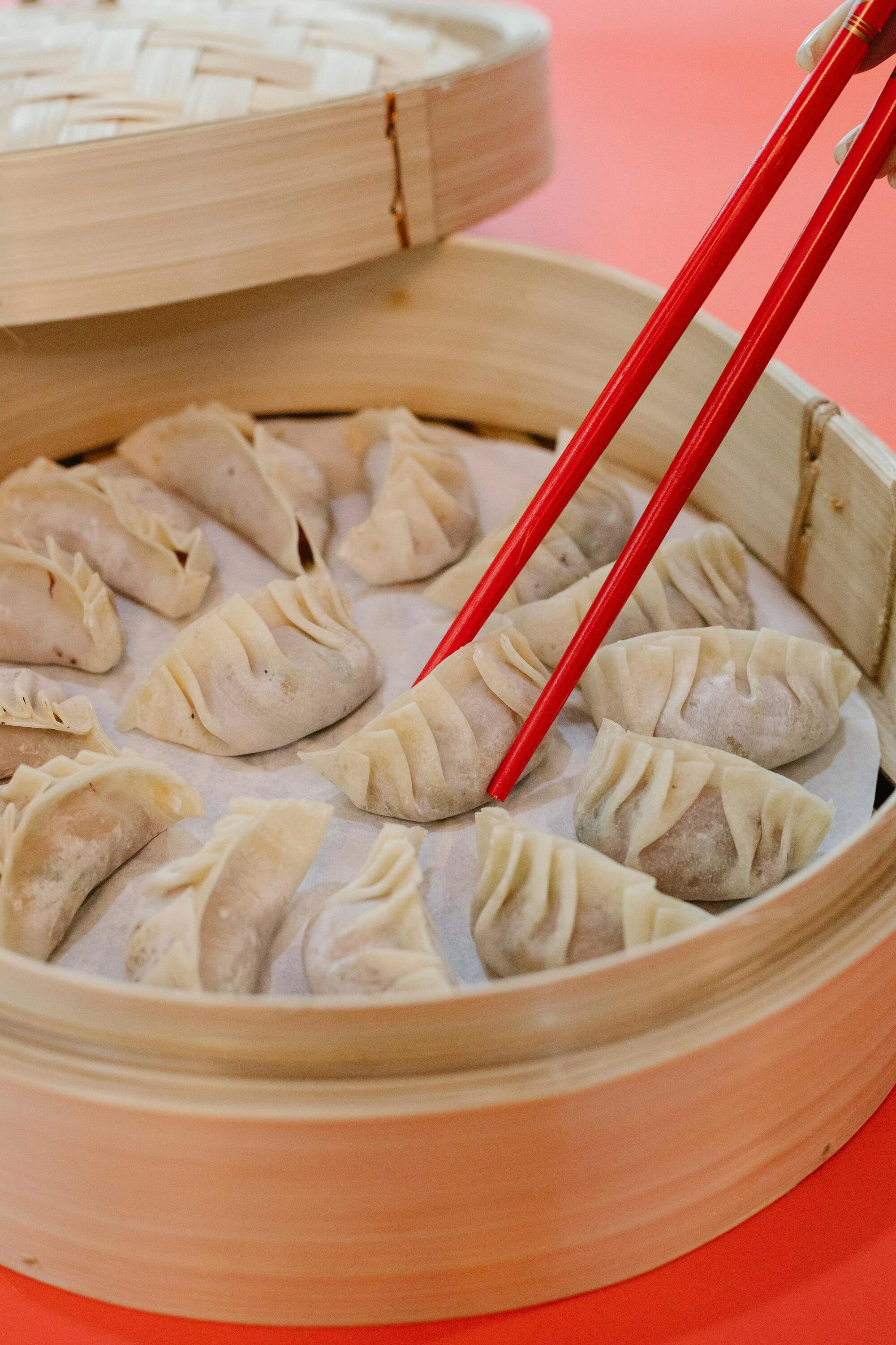 Red chopsticks pick up a gyoza dumpling from a wooden steamer basket