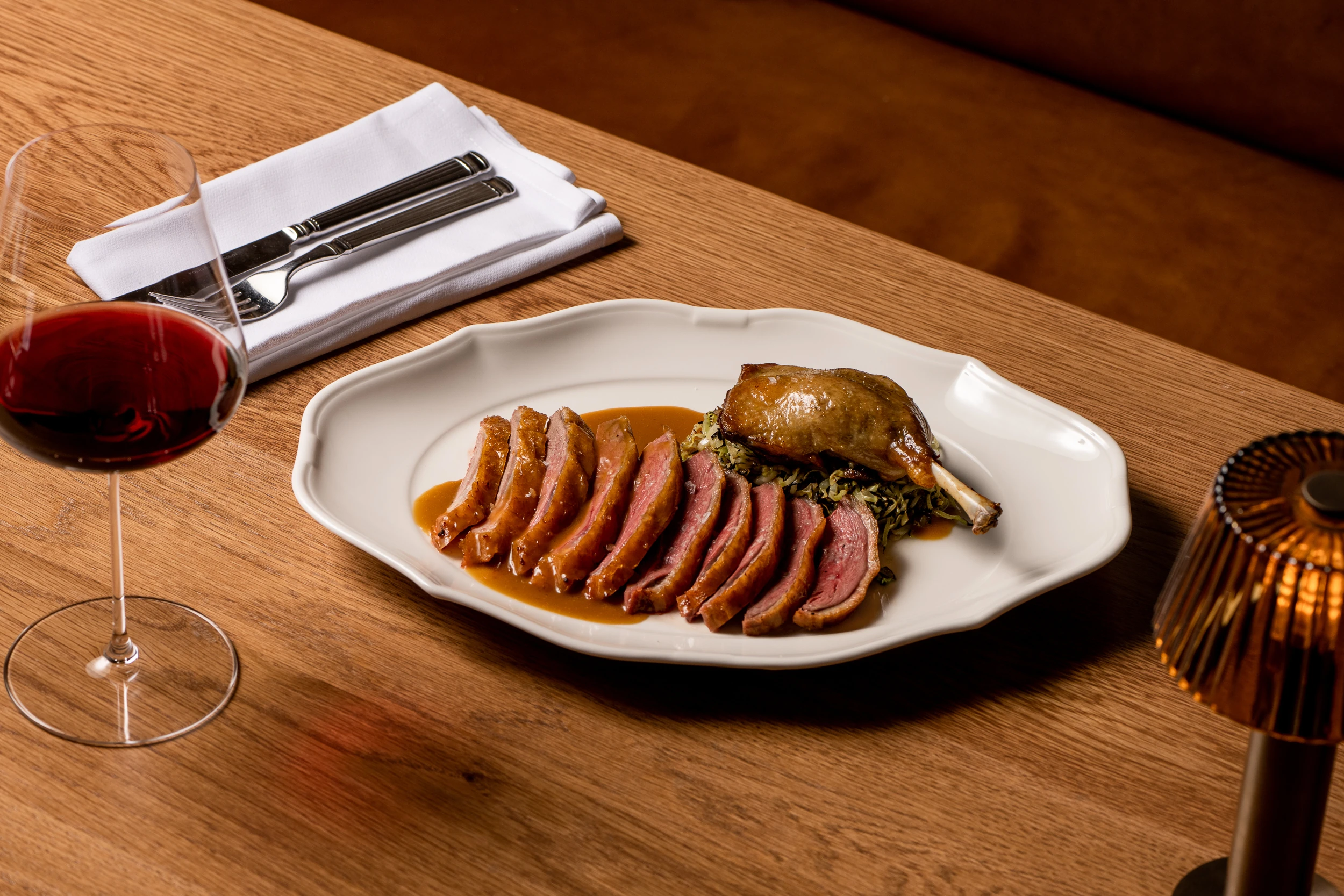 A plate of Great Ocean duck, sugarloaf cabbage and jus on a wooden table with cutlery, a lamp and a glass of red wine.