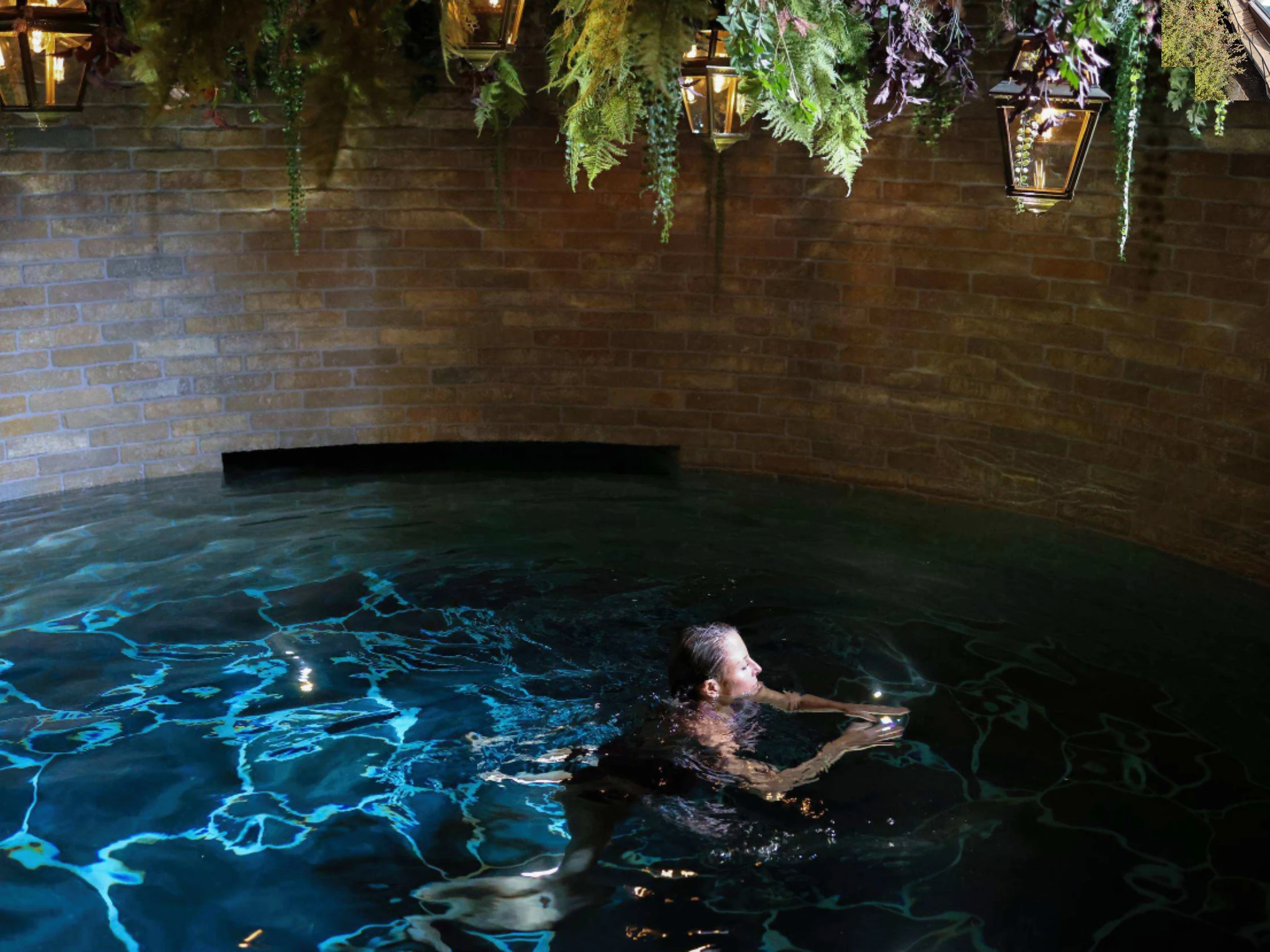 A woman relaxing in an indoor brick pool illuminated by hanging lanterns and lush ceiling foliage at a spa in Lidingö