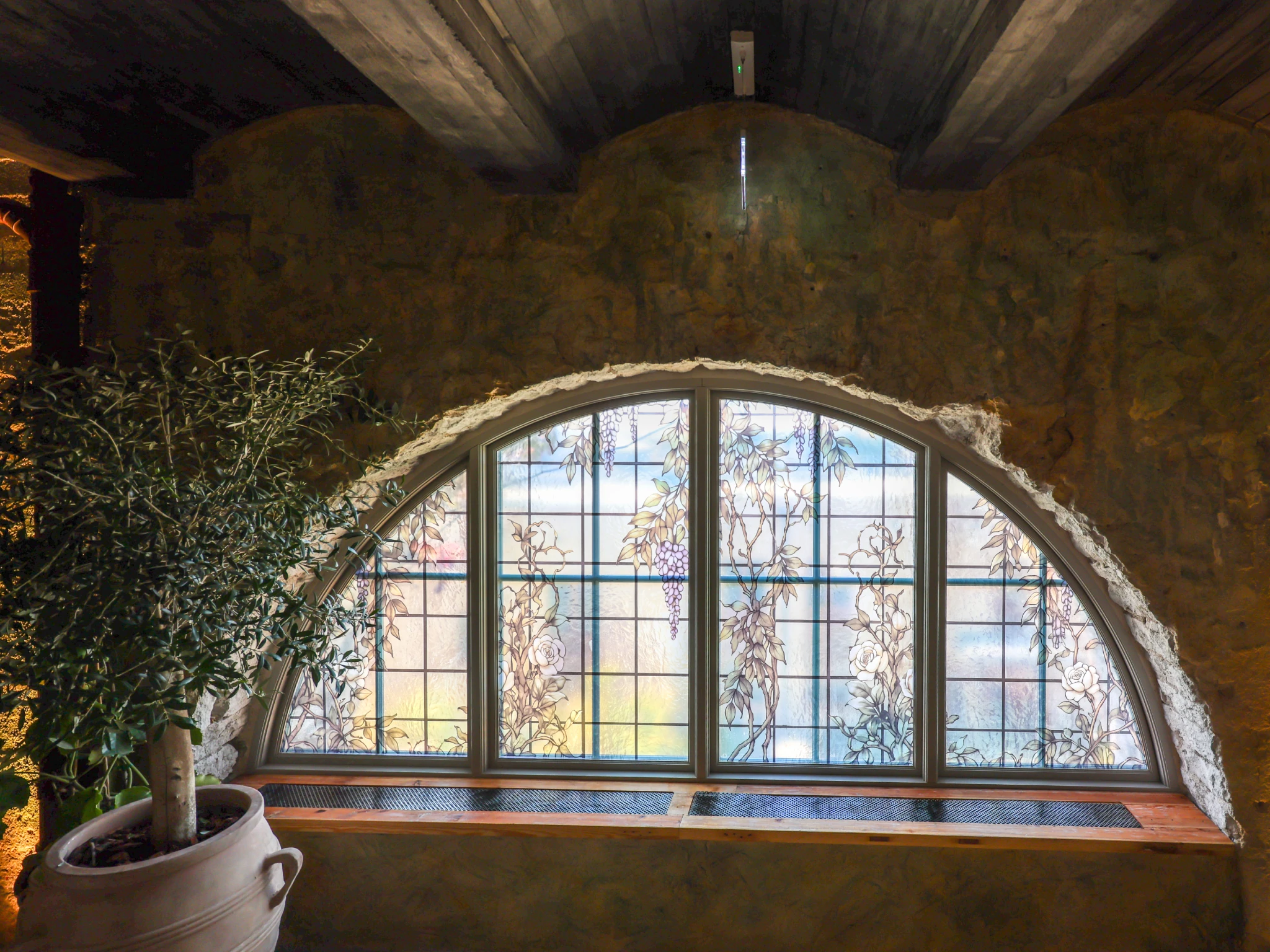 An arched stained glass window with vine motifs set into a rustic stone wall next to a potted olive tree at Villa Foresta.