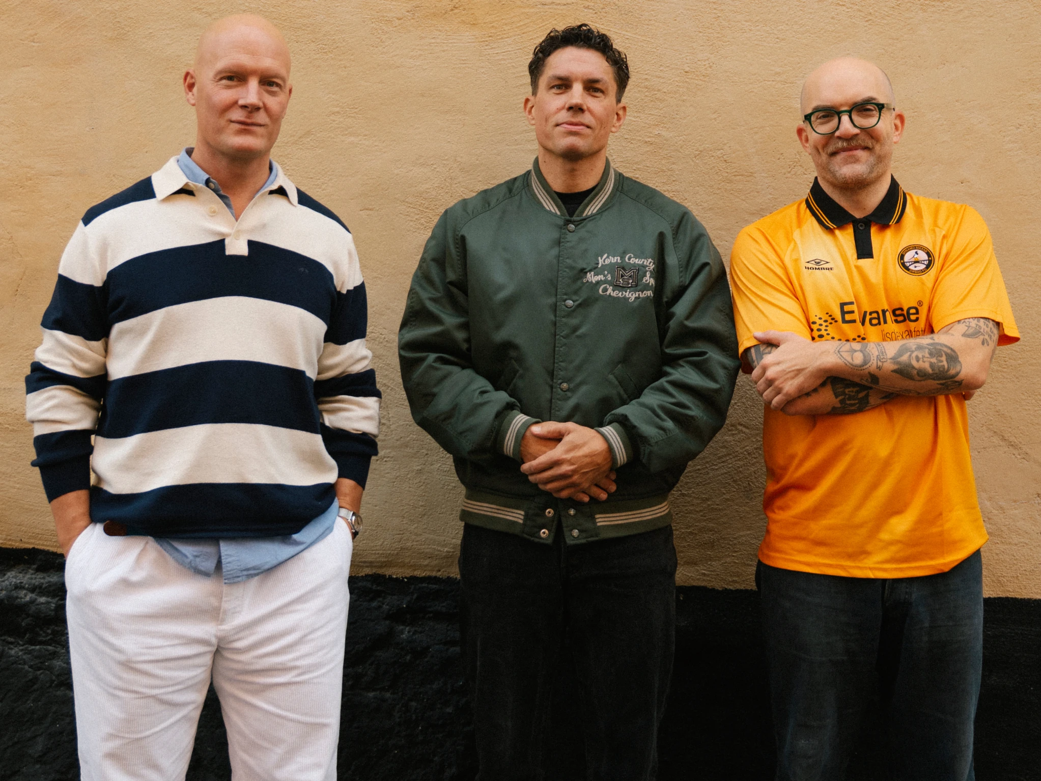 Three men against an ochre wall, one in a striped rugby shirt, one in a green varsity jacket and one in an orange football shirt.