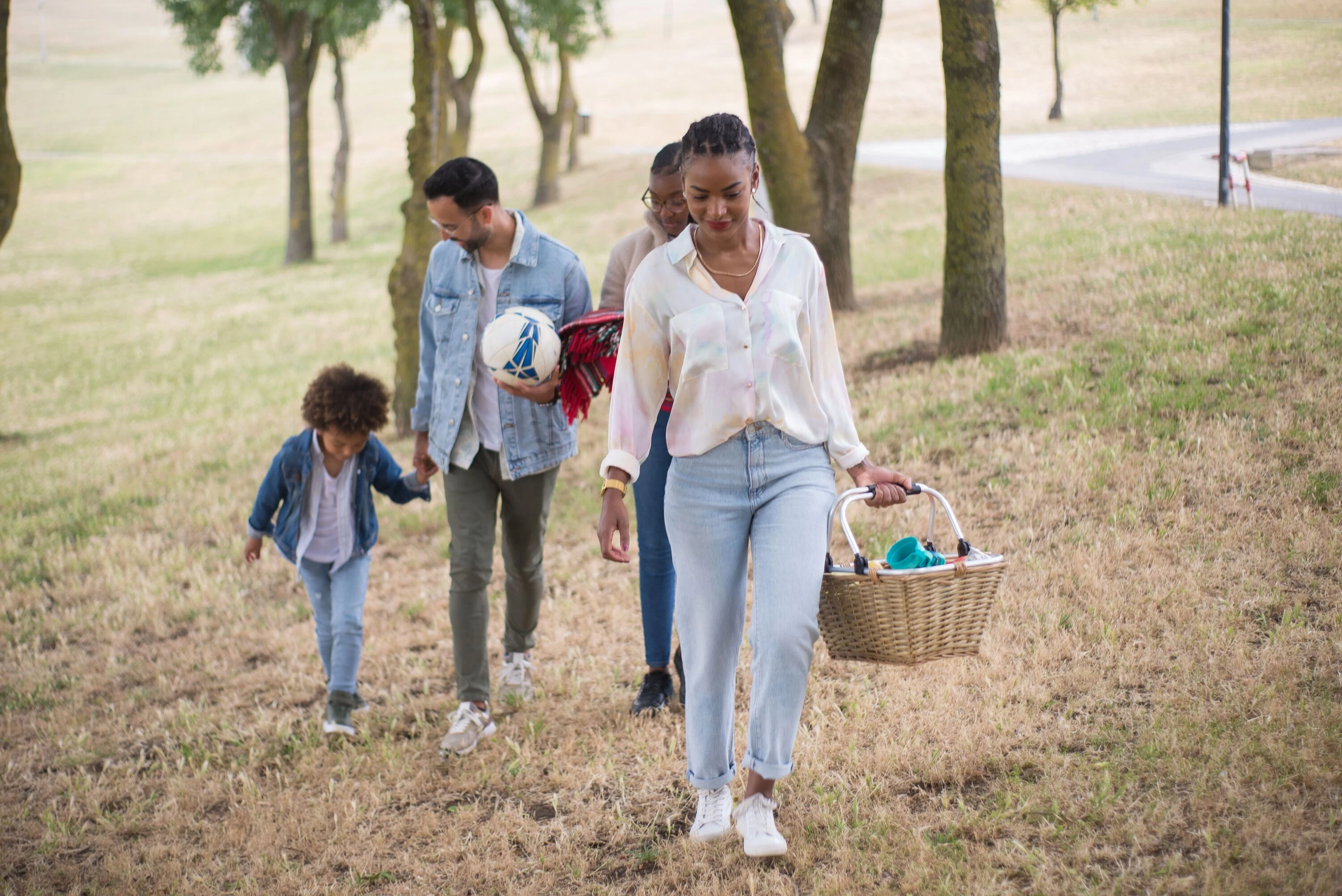 A joyful family walking in a park, carrying a picnic basket and a ball on a sunny day.