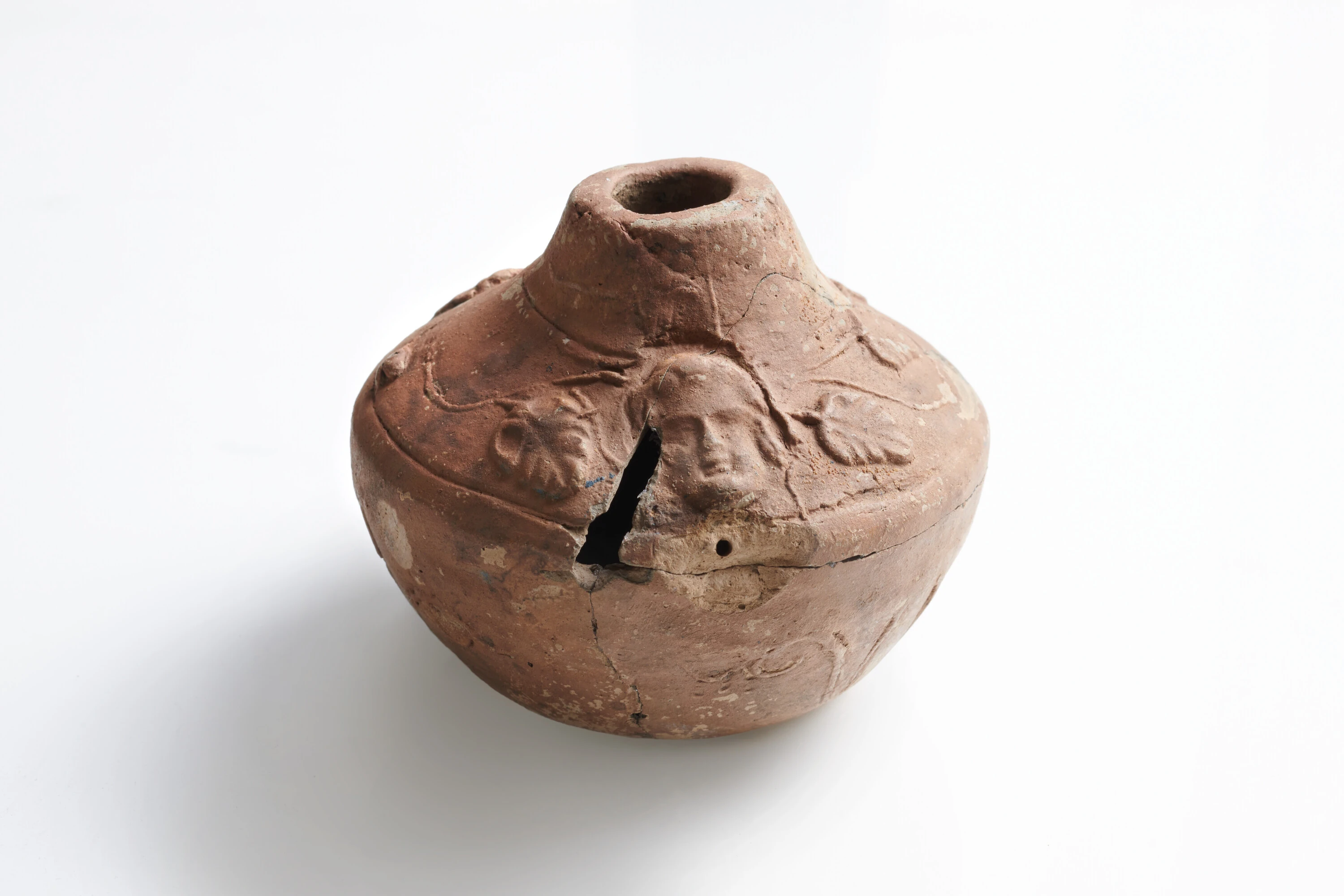 A cracked, round, ancient Roman clay vessel with a narrow neck and faded decorative patterns, shown on a plain white background