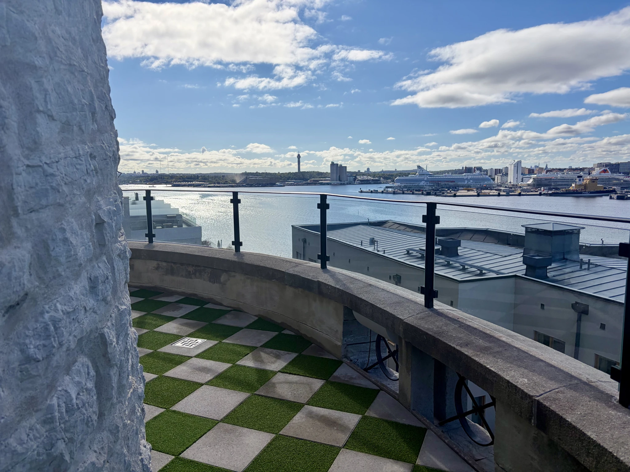A outdoor balcony terrace featuring a checkered green and grey tiled floor and a glass railing, looking out over the water toward Stockholm city, cruise ships, and a partly cloudy sky at Aqua di Foresta.