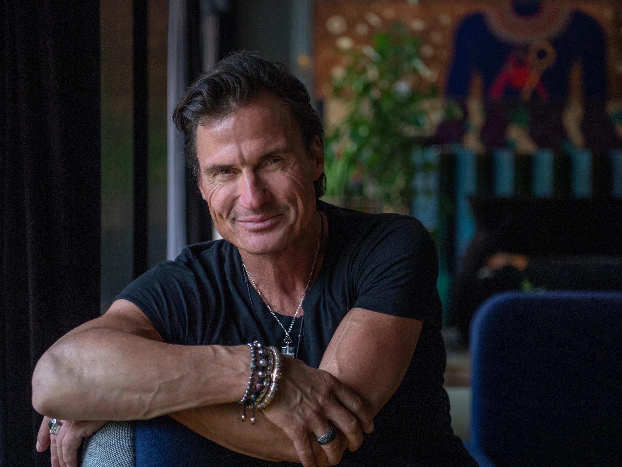 Petter Stordalen, owner of Strawberry, smiling in a black T-shirt and stacked bracelets, leaning on the back of a blue armchair in a dimly lit room.