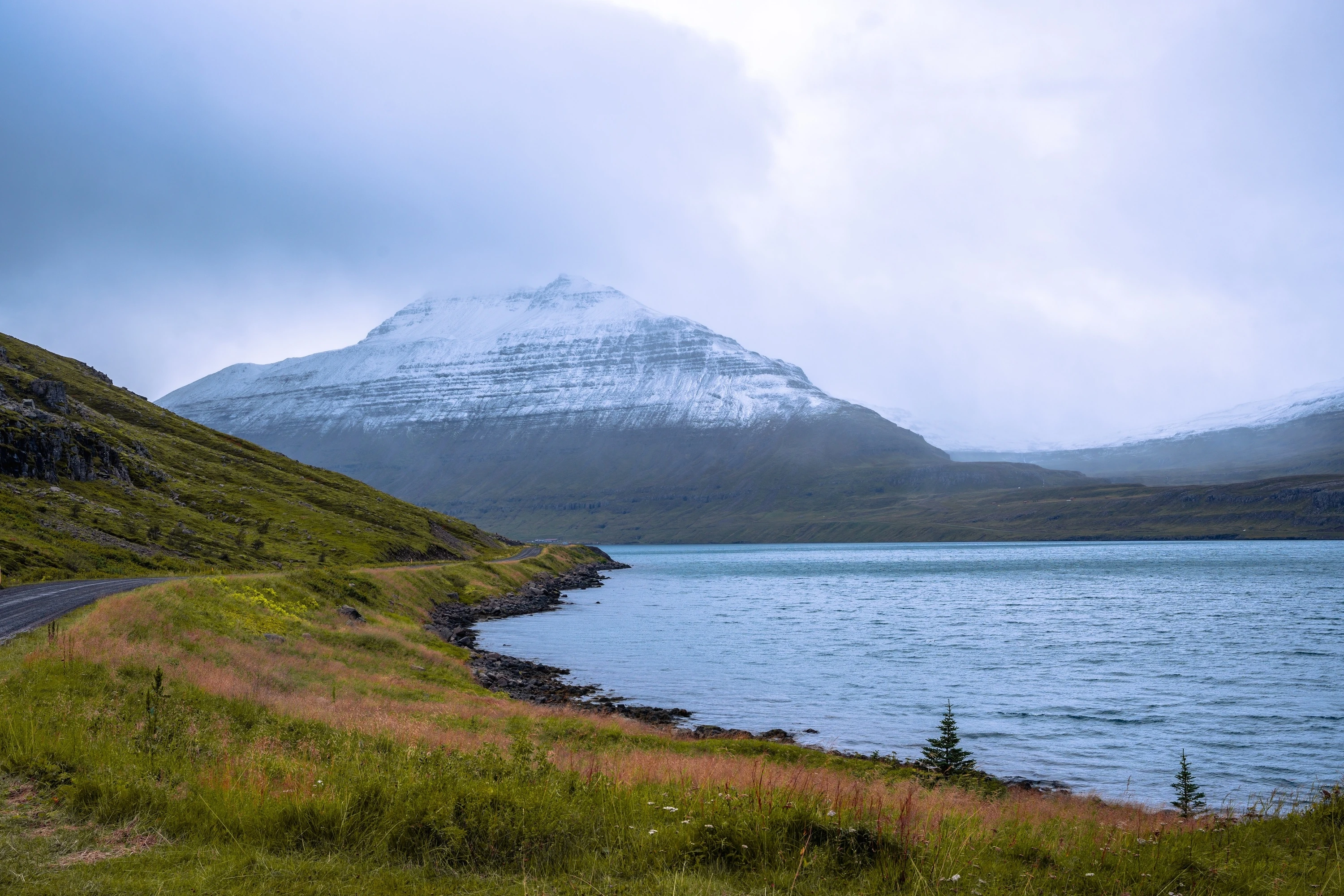 A winding road on the side of a lake with a snowy mountain in the background