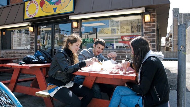 Three people eating on a picnic table outside the Wieners Circle.