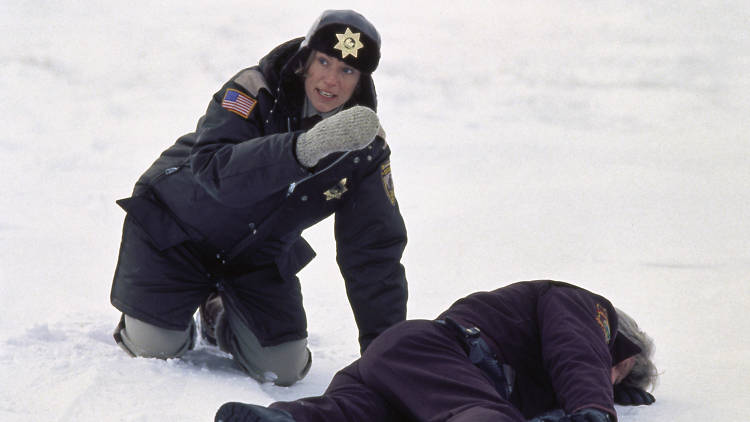 Frances McDormand in Fargo cop unifmorm, crouching in snow over a slumped body