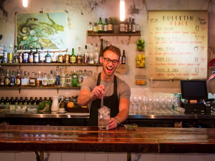 A bartender smiling behind the bar at Bulletin Place stiring a c