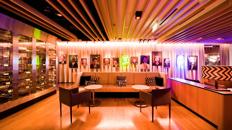 A shot of the lobby area at QT Canberra with a couch, a large wine cabinet and portrait posters of famous people hung on the wall