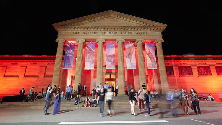 Picture of the Art Gallery of New South Wales at night from across the road, with the building lit up with red lights, and banners for Sydney Moderns exhibition hanging from the front.