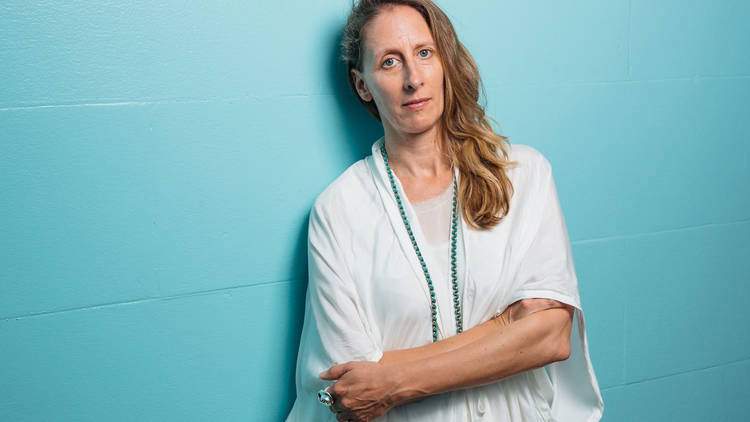Image of Biennale of Sydney curator Stephanie Rosenthal with her hair out and wearing loose-fitting white robes, standing with her arms folded against the bright turquoise wall of Mortuary Station