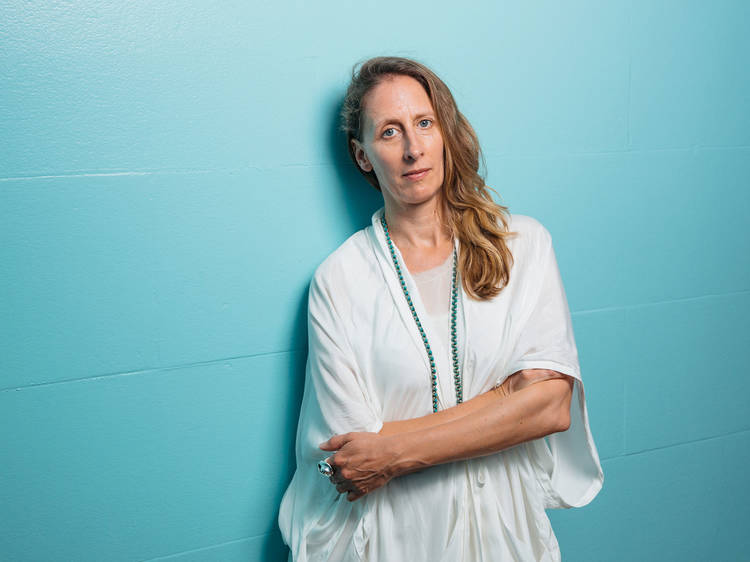 Image of Biennale of Sydney curator Stephanie Rosenthal with her hair out and wearing loose-fitting white robes, standing with her arms folded against the bright turquoise wall of Mortuary Station