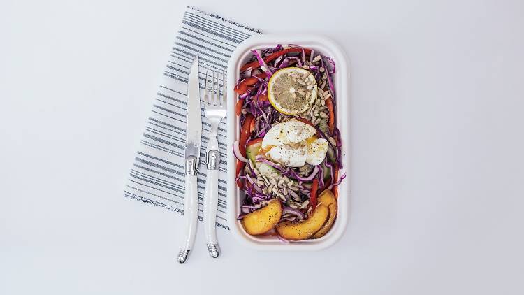 Takeaway box filled with cabbage salad, peach slices, sunflower seeds and a poached eggs