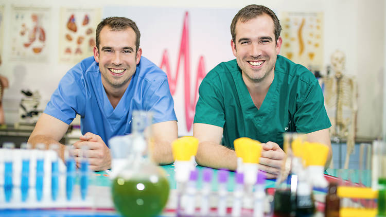 Two men standing at a science lab table of experiments for a kids show.
