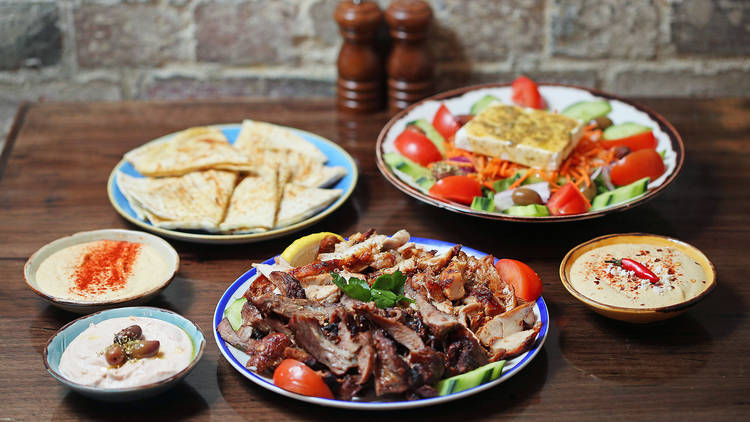 On a wooden table there is a plate of souvlaki meat, pita bread, a greek salad and bowls of dips