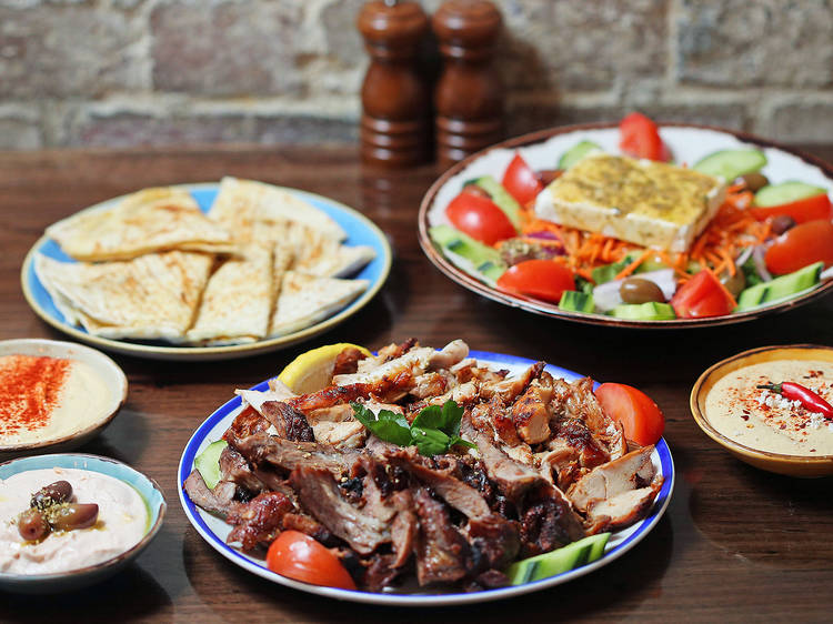 On a wooden table there is a plate of souvlaki meat, pita bread, a greek salad and bowls of dips