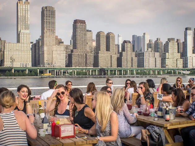 A few dozen people eat at picnic tables with a view of the river and skyline.