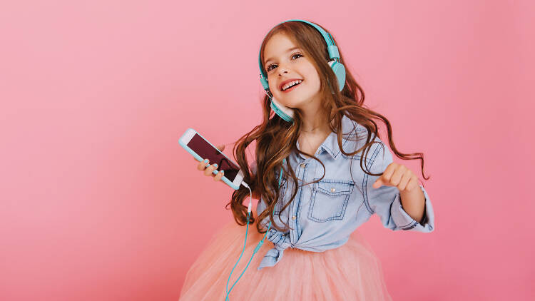 Little girl with long hair holding onto an iPhone with green headphones dancing while wearing a pink tutu and denim shirt while standing in front of a pink wall