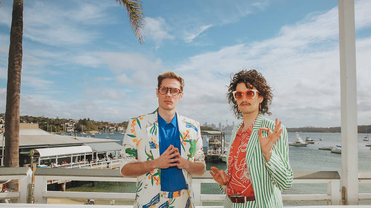 The band members stand on a deck in front of the beach.