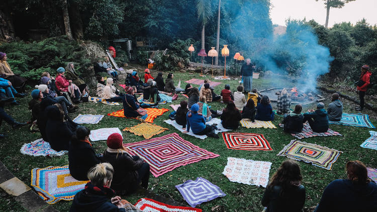 People sit on colourful rugs in the forest.