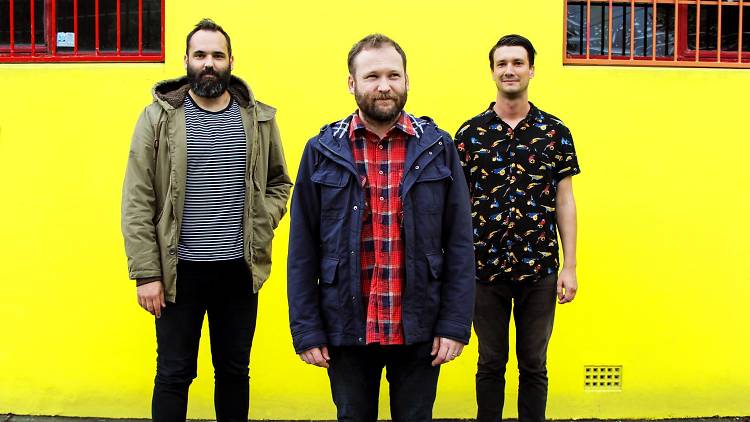 Band members stand in front of a yellow wall.