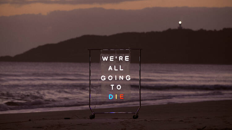 We're All Going to Die neon sign on a beach