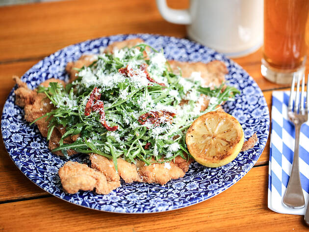 A chicken schnitzel on a plate topped with salad, cheese and a lemon