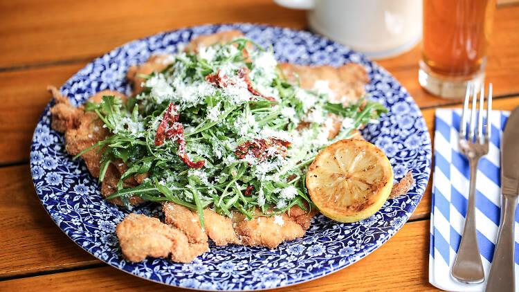 A chicken schnitzel on a plate topped with salad, cheese and a lemon