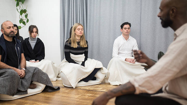 Four people with eyes closed sitting cross legged practicing meditation lead by Manoj Dias