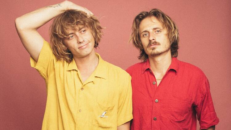 Band members of Lime Cordiale standing in front of a burnt orange wall. The band member on the left is wearing a bright yellow shirt and the band member on the right is wearing a red shirt