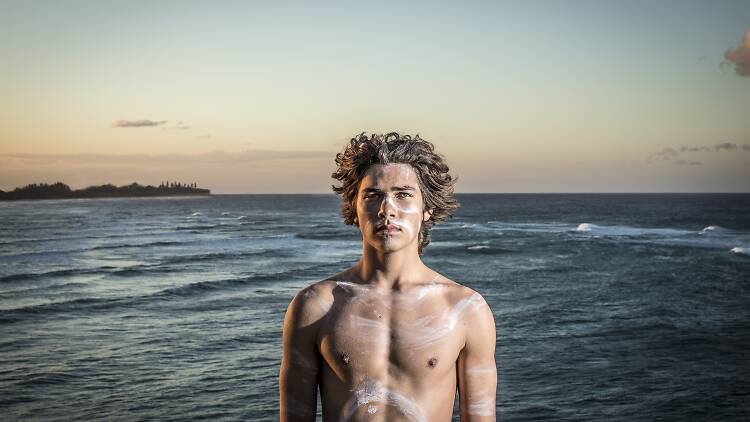 Press image for 'The Spirit of Churaki'; young man standing facing the camera with ocean backdrop, representing the true story of Churaki, a Coodjingburra man of the Bundjalung tribe, heralded as the Gold Coast’s first surf lifesaver 