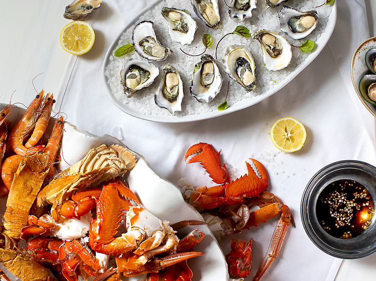 A birdseye view of a table with white table cloth and plates with lobsters, crabs, balmain bugs, oysters on ice, mussels and lemon halves, with dipping sauce