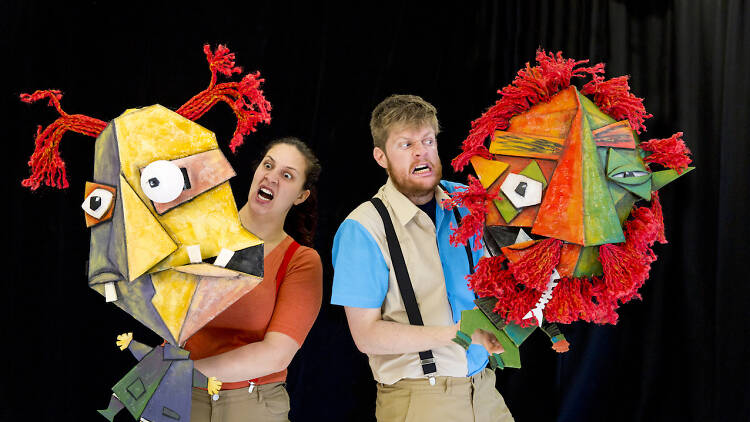 Two actors holding colourful puppets in front of a black wall