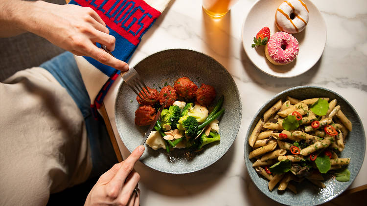 A plate of meatballs being cut into next to a plate of pasta and a plate of doughnuts