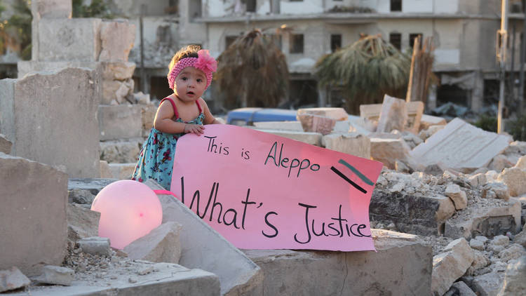A small child holds a sing reading 'This is Aleppo - What's Justice' while standing in a crumbling building.