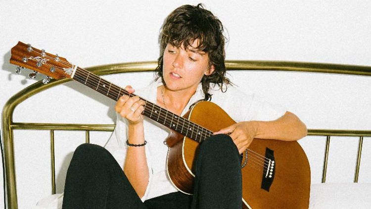 Press shot of Courtney Barnett playing a guitar on a bed