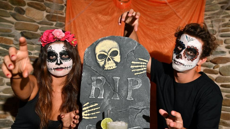 Two people in skull makeup leer at the camera, a headstone and a cocktail sit between them.