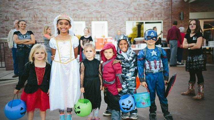 A row of children in halloween costumes smile at the camera, they are holding plastic pumpkins full of treats.
