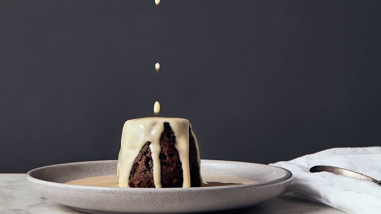 A Christmas pudding sits on a large white plate with custard being dripped on to it
