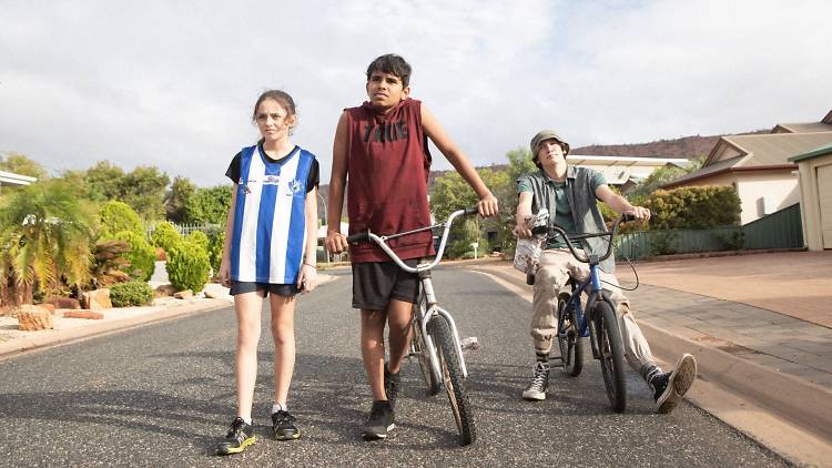 Three children stand on a suburban street looking off into the distance, two are on bikes.