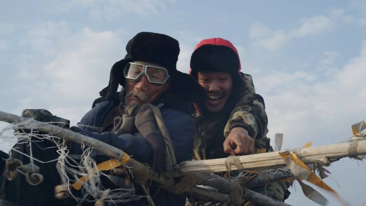 A young man supports an elderly man on a makeshift sled, they are both wearing warm clothes and hats.