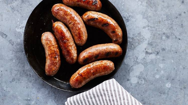 A pan of thick juicy sausages on a grey bench with a striped tea towel wrapped around the handle.