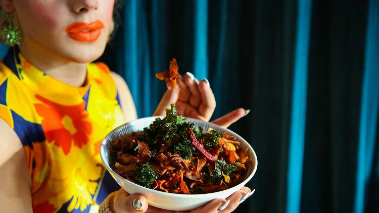 A person with bright makeup, orange lipstick and a floral dress holds up a salad full of sweet potato crisps.