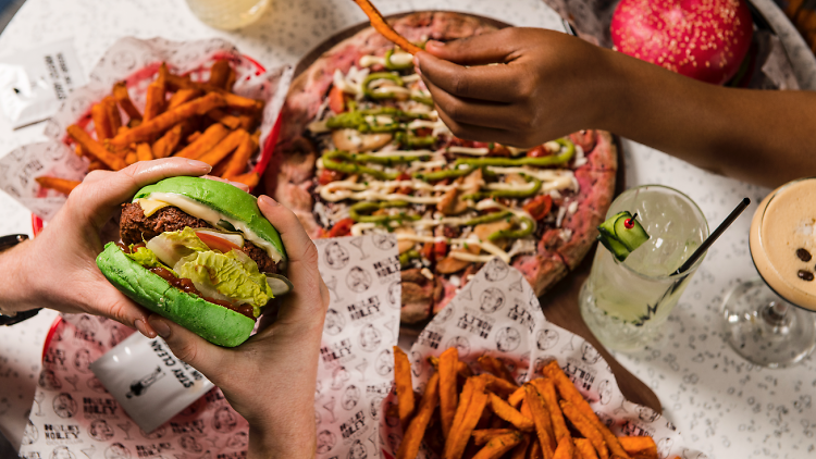 A table of food including fries and pizza, someone holds a burger with a bright green bun in the foreground.
