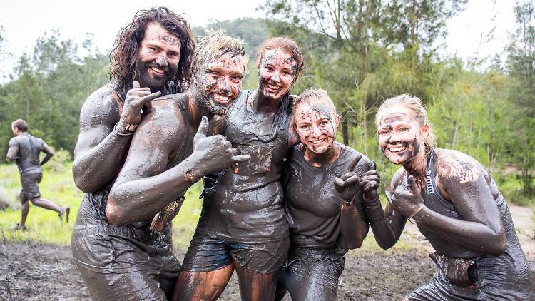 A group of five people covered in mud are smiling, embracing each other, and giving thumbs up.