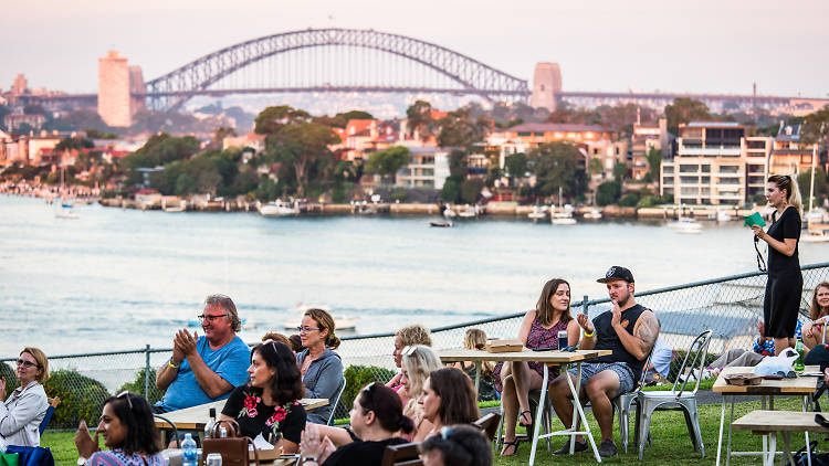 People sitting at tables on Cockatoo Island with the Sydney Harbour Bridge in the background.