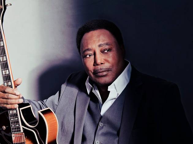 George Benson George Benson poses with his guitar in a grey suit.