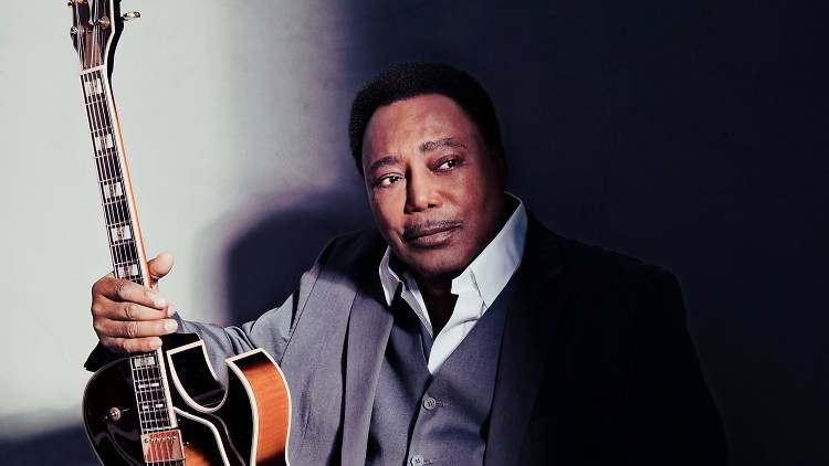 George Benson poses with his guitar in a grey suit.