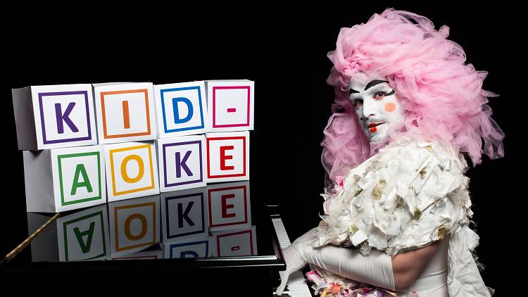 A performer in elaborate pink wig and white makeup plays a grand piano.